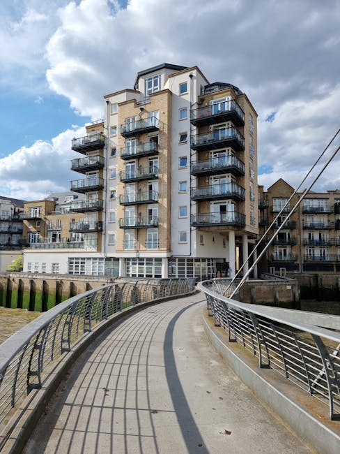 A modern multi-story residential building with a light-colored facade and numerous glass balconies, situated along a waterfront promenade. The promenade features a curved pathway with metal railings o