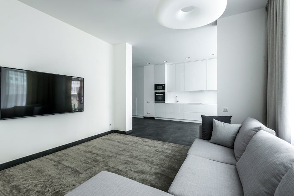 A modern multi-story residential building with a light-colored facade and numerous glass balconies, situated along a waterfront promenade. The promenade features a curved pathway with metal railings o