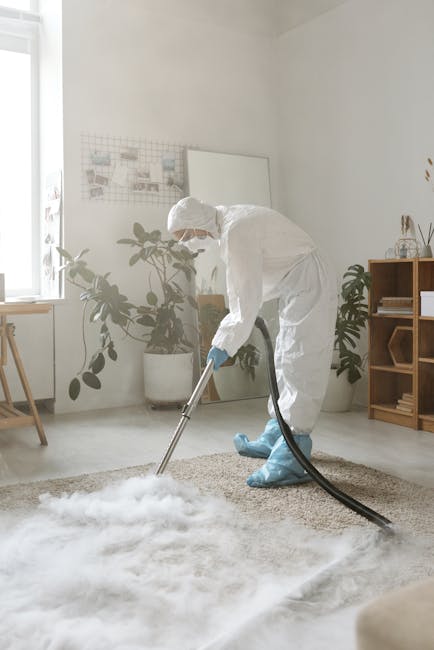A professional cleaner dressed in a white protective suit, face mask, and blue shoe covers is using a wet vacuum cleaner to deep clean a beige carpet in a well-lit living room. The room features white walls, wooden furniture, and potted plants near a window, with natural light illuminating the space. The cleaner's focused posture suggests thorough surface cleaning and sanitisation, characteristic of Bermondsey Carpet Cleaning's same-day emergency carpet cleaning services in Bermondsey (SE1). The scene highlights the use of modern cleaning equipment to achieve high standards of hygiene and carpet maintenance, ensuring a fresh and pristine appearance.