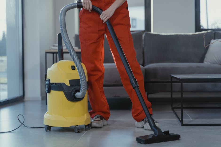 A professional cleaner from Bermondsey Carpet Cleaning is performing deep surface cleaning on a beige, textured carpet in a well-lit, modern living room. The cleaner is dressed in full protective gear, including a white coverall, face mask, gloves, and shoe covers, and is using a steam cleaning machine with a black hose. The room features a wooden bookshelf with decor items, a large mirror leaning against the wall, and potted indoor plants. The flooring appears clean and damp from the cleaning process, highlighting the sanitation and thoroughness of the service provided by Bermondsey Carpet Cleaning for emergency same-day carpet cleaning in Bermondsey (SE1).