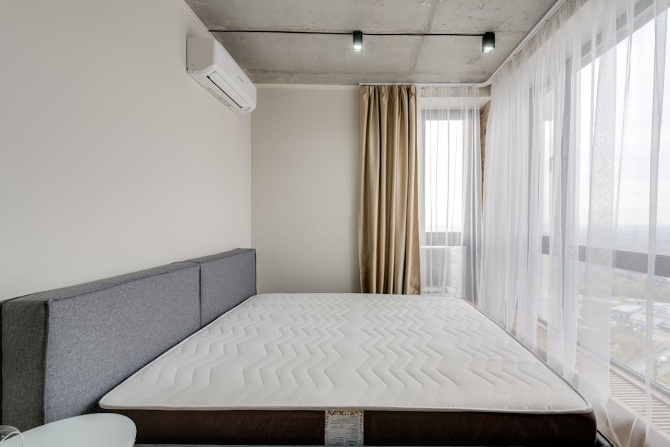 A modern bedroom featuring a double bed with a dark grey upholstered headboard and a white mattress, situated near large floor-to-ceiling windows dressed in sheer white curtains and a beige blackout curtain. The room has a minimalist design with plain white walls, a mounted air conditioning unit, and natural light flooding in, highlighting the clean, dust-free surfaces. The light grey carpeted floor and the polished window frames are visible, emphasizing the room’s tidy and hygienic appearance suitable for surface cleaning and sanitisation. Bermondsey Carpet Cleaning promotes thorough domestic cleaning practices within such spaces to meet local Southwark Council rules for mattress sanitation.