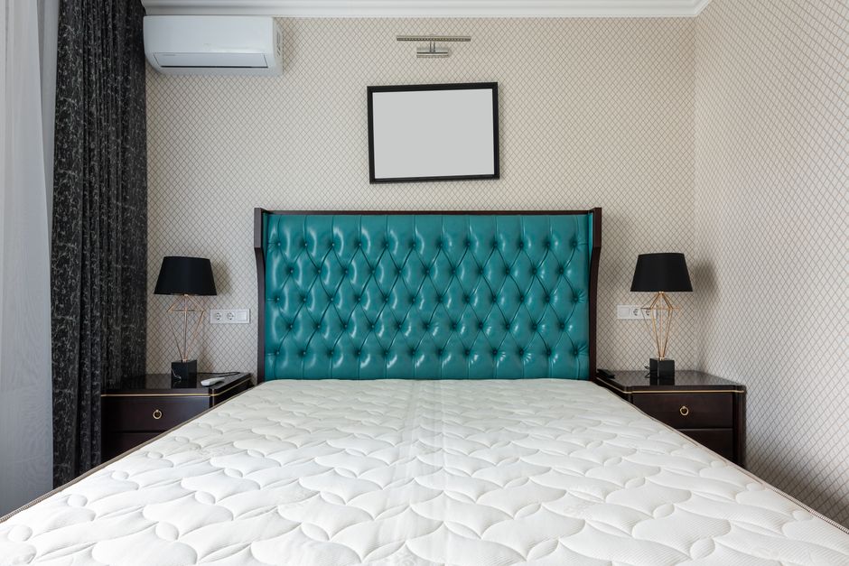 A well-maintained bedroom features a large bed with a bright turquoise, diamond-stitched upholstered headboard, positioned centrally against a wallpapered wall with a small, blank rectangular picture frame above. On either side of the bed are dark wood nightstands with gold accents, each holding black table lamps with gold geometric bases. The room has a white air conditioning unit mounted high on the wall, and dark curtains partially cover a window on the left. The mattress appears clean and hygienic, suggesting professional surface cleaning and sanitisation, consistent with domestic cleaning standards promoted by Bermondsey Carpet Cleaning. The neutral interior, with subtle lighting and orderly arrangement, emphasizes cleanliness and a well-kept environment.