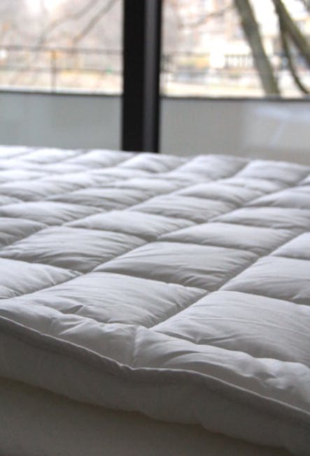 A modern bedroom featuring a double bed with a dark grey upholstered headboard and a white mattress, situated near large floor-to-ceiling windows dressed in sheer white curtains and a beige blackout curtain. The room has a minimalist design with plain white walls, a mounted air conditioning unit, and natural light flooding in, highlighting the clean, dust-free surfaces. The light grey carpeted floor and the polished window frames are visible, emphasizing the room’s tidy and hygienic appearance suitable for surface cleaning and sanitisation. Bermondsey Carpet Cleaning promotes thorough domestic cleaning practices within such spaces to meet local Southwark Council rules for mattress sanitation.